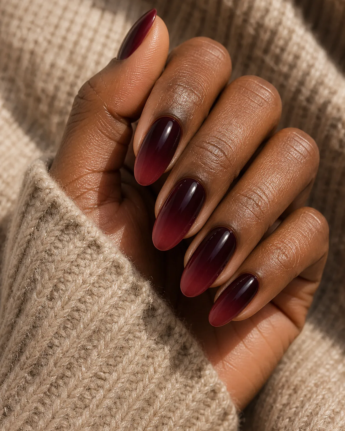 Close-up of oval nails in a wine-to-burgundy deep ombre, the blend rich and same-family, on deep skin resting on a soft knit sleeve