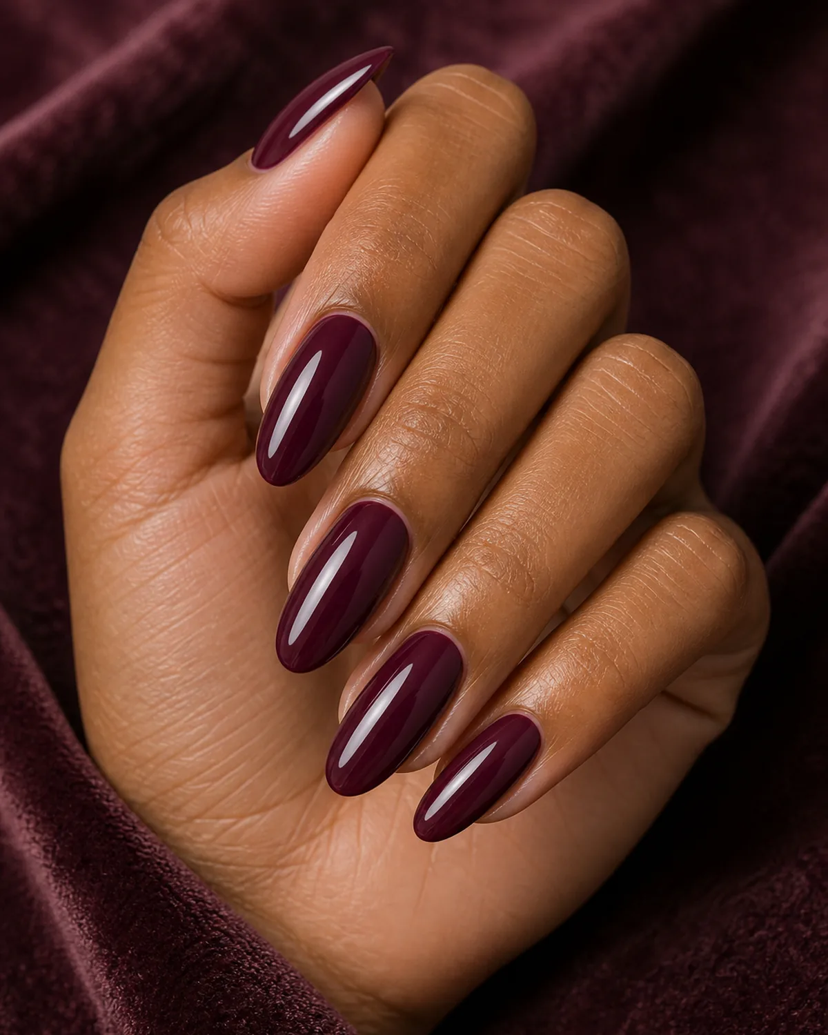 Close-up of long oval nails in deep burgundy plum polish covering the entire nail with both side walls curving inward to a soft even U-shaped free edge, on medium-deep skin with hand tilted pinky down on plum velvet
