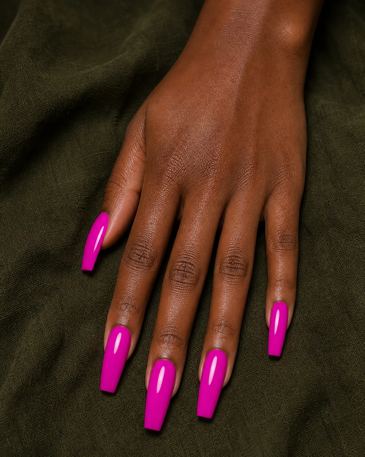 Close-up of long coffin nails in vivid hot magenta pink gloss covering the entire nail surface in an electric pink-violet read, on deep skin closed into a soft fist on dark mossy linen