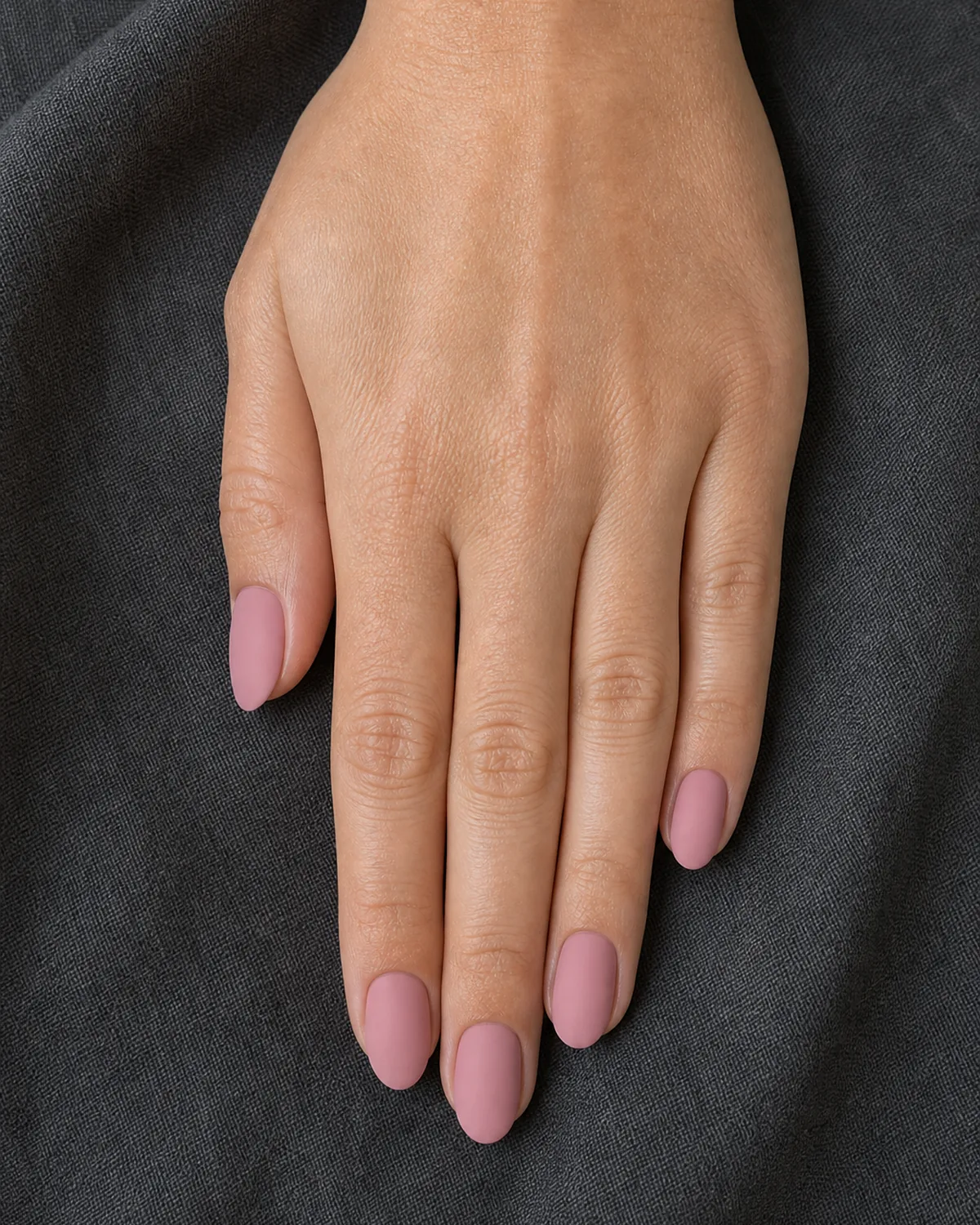 Close-up of medium oval nails in muted dusty pink with a flat matte velvet finish covering the entire nail, on light-medium skin resting on charcoal linen