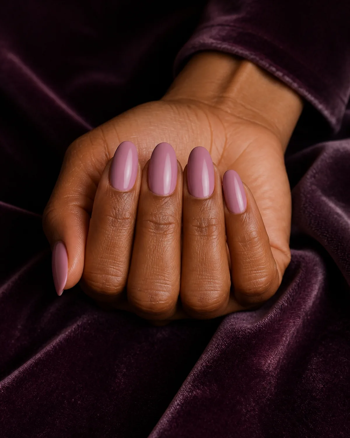 Close-up of medium almond nails in cool mauve-pink cream lacquer with a soft violet-pink lean covering the entire nail surface, on medium-deep skin against plum velvet