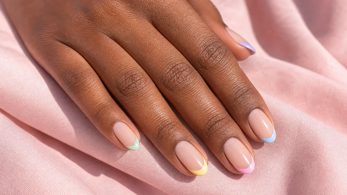 Close-up of short-to-medium almond nails in a pastel French, a sheer base with thin crisp tips each in a different soft pastel, on deep skin against soft-pink fabric