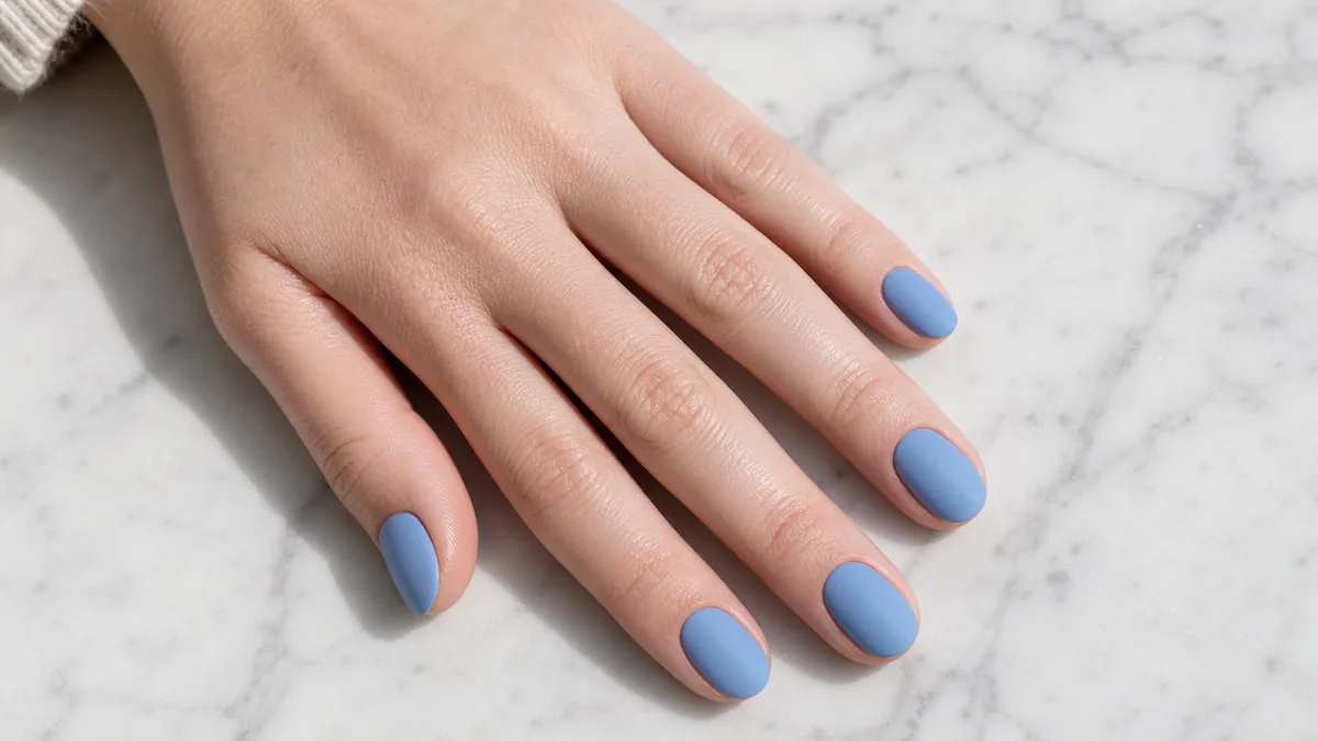 Top-down macro of short, rounded nails in a powdery cornflower blue on fair skin resting on pale marble