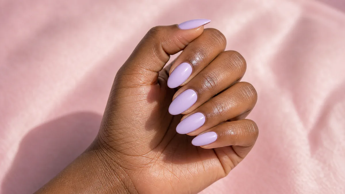 Close-up of almond nails in a milky soft lilac, fingers gently curled, on deep skin against a soft-pink background