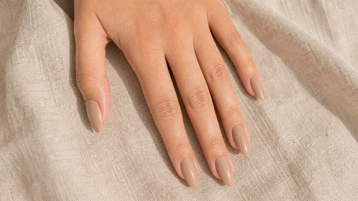 Top-down close-up of almond nails in a creamy, warm latte nude on light-medium skin, the hand flat on oat linen