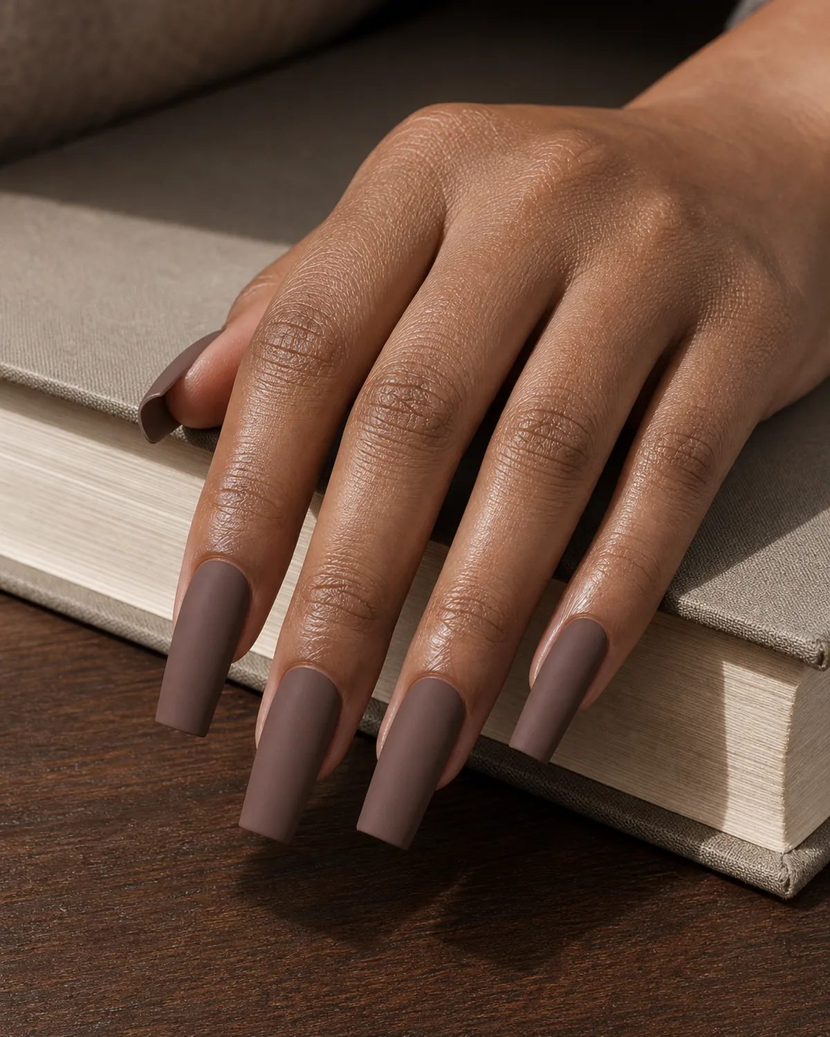Close-up of long square nails in a matte warm mocha brown polish covering the entire nail with both side walls perfectly straight and parallel, free edge flat horizontal and tip corners sharp at 90 degrees, on medium-deep skin with hand flat and fingers pressed tightly together on dark walnut wood