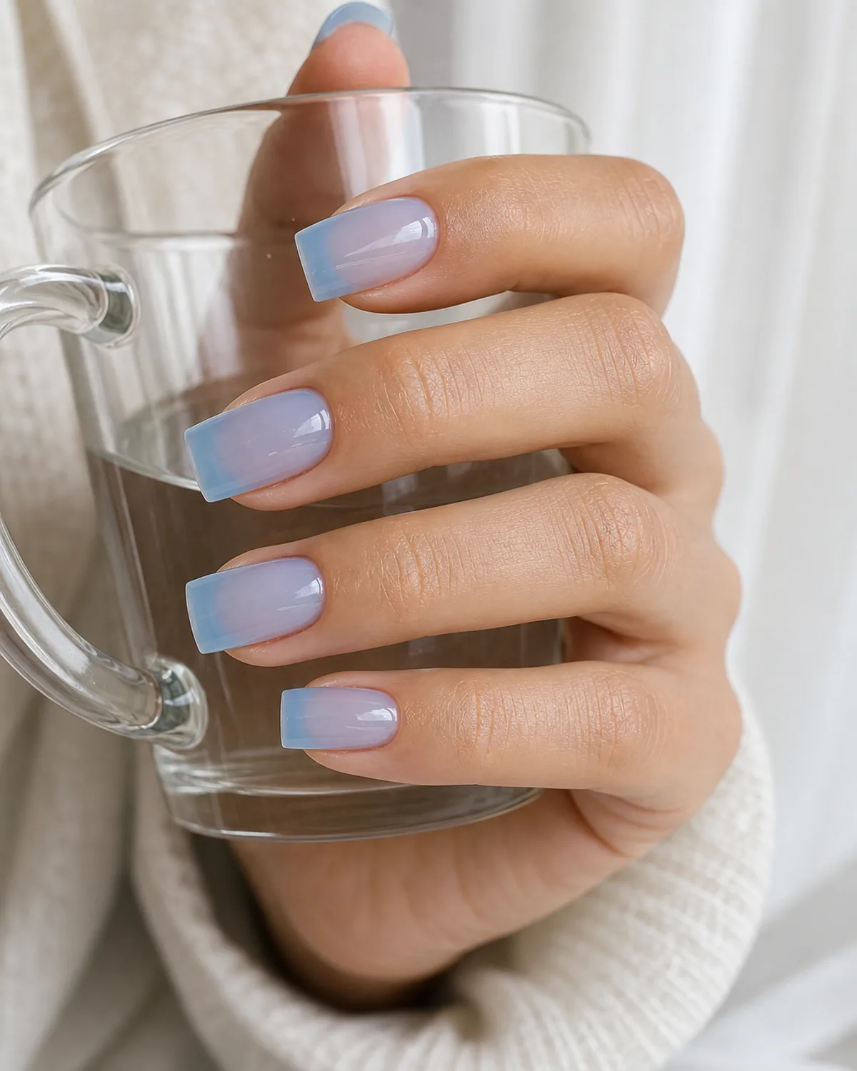 Close-up of medium square nails in a cool sky-blue jelly polish covering the entire nail with both side walls perfectly straight and parallel, free edge flat horizontal and tip corners sharp at 90 degrees, on light-medium skin with hand three-quarter view and fingers in a loose curl holding a glass mug