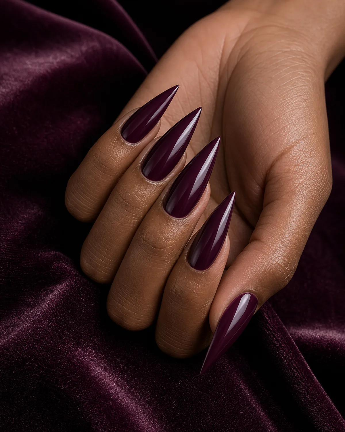 Close-up of long stiletto nails in deep burgundy plum polish covering the entire nail with both side walls filed inward in a dramatic steep taper converging at the tip into a single sharp acute talon point, on medium-deep skin with hand tilted pinky down on plum velvet