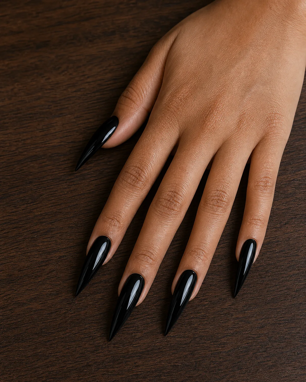 Close-up of long stiletto nails in glossy jet-black polish covering the entire nail with both side walls filed inward in a dramatic steep taper converging at the tip into a single sharp acute talon point, on tan skin with hand flat thumb tucked and fingers splayed on dark walnut wood