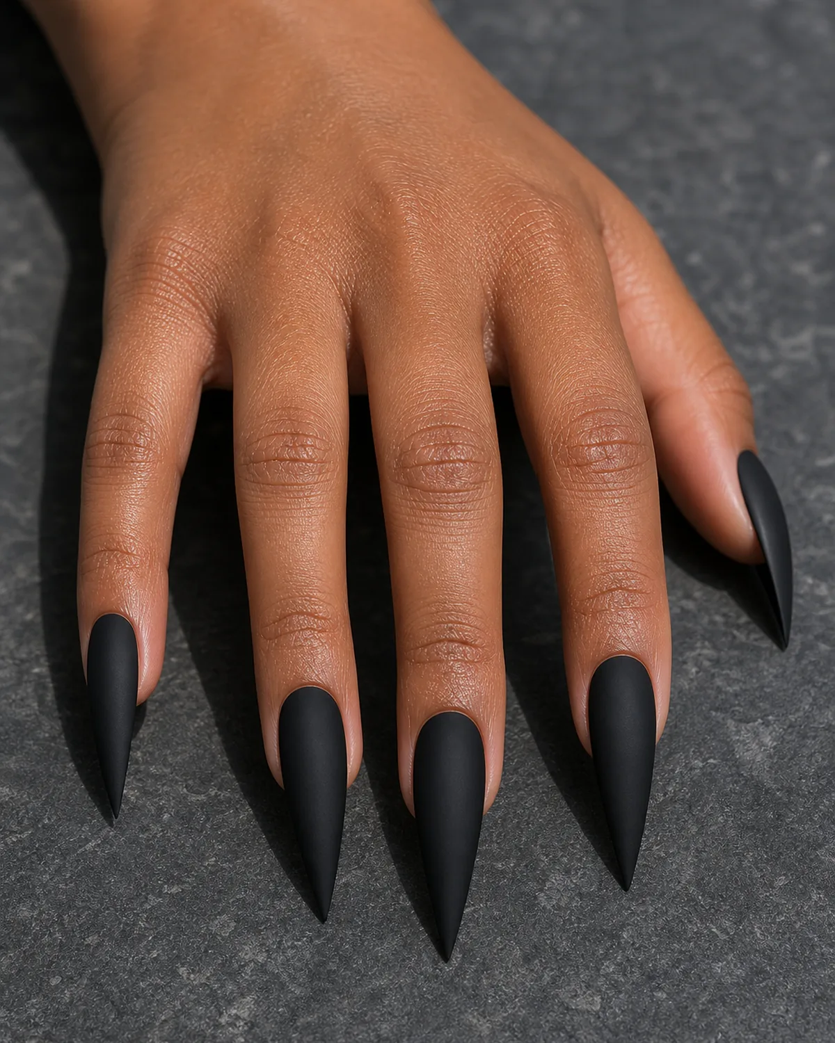 Close-up of long stiletto nails in matte black polish covering the entire nail with both side walls filed inward in a dramatic steep taper converging at the tip into a single sharp acute talon point, on medium-deep skin with hand arched fingers down fanned piano-style on charcoal stone