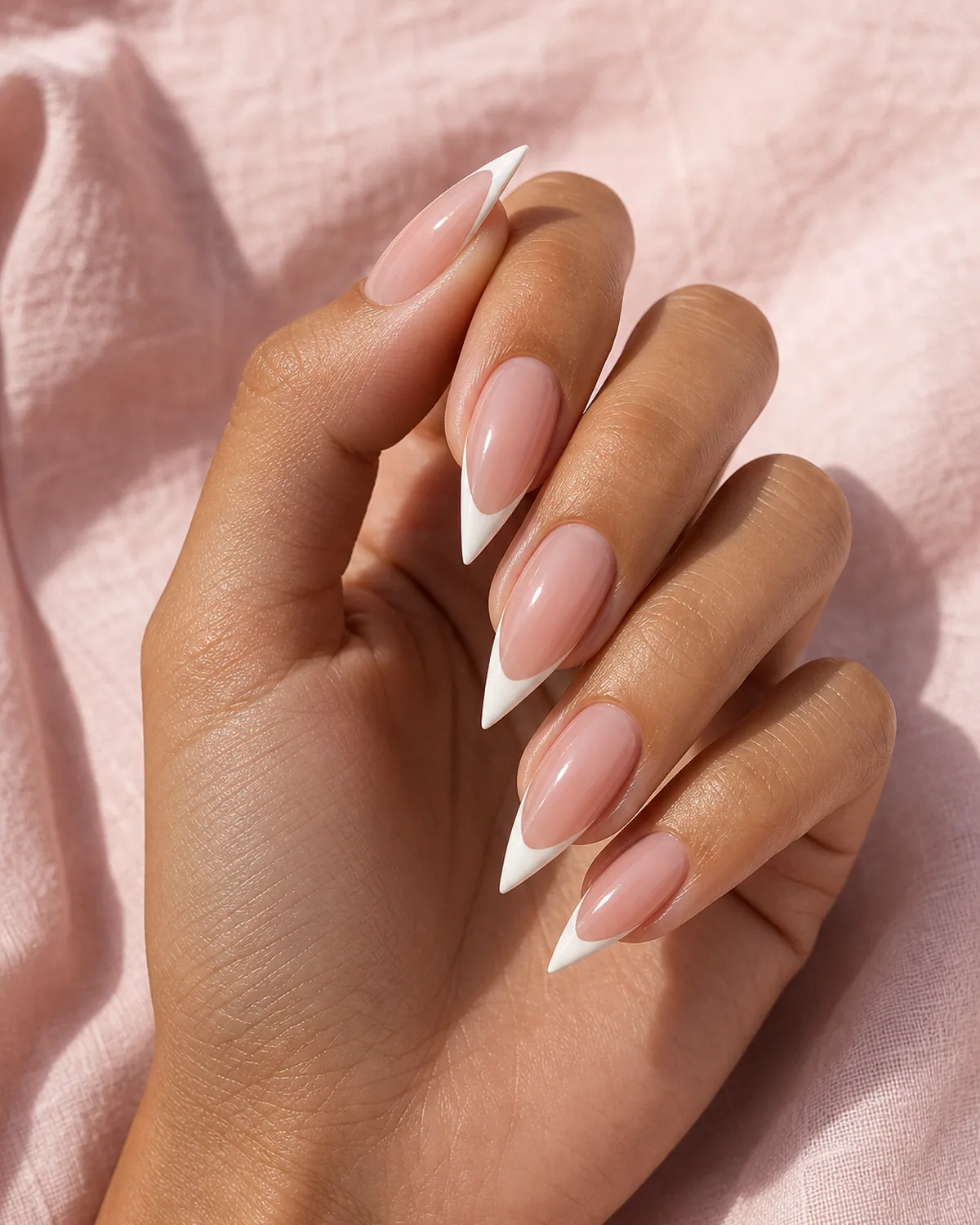Close-up of medium stiletto nails wearing a classic French manicure with a thin warm-white smile band that follows the dramatic inward taper and closes into the single sharp acute talon tip, on medium skin with fingertips touching thumb in a soft pinch gesture on soft-pink linen