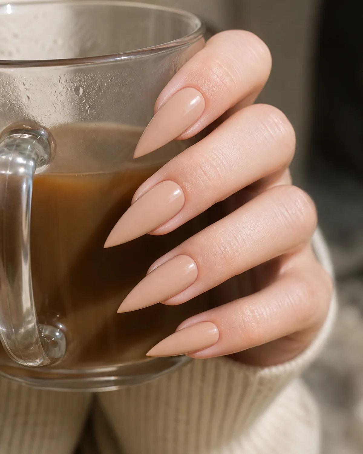 Close-up of medium stiletto nails in opaque nude beige polish covering the entire nail with both side walls filed inward in a dramatic steep taper converging at the tip into a single sharp acute talon point, on fair skin with three-quarter hand view and fingers in a loose curl around a glass mug
