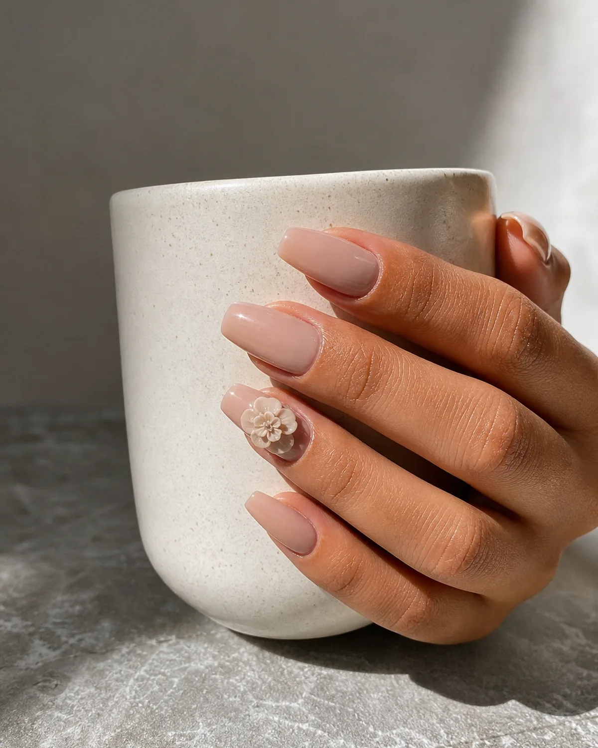 Close-up of medium coffin nails with a nude base and a small sculpted 3D flower built from layered petals, on medium skin cradling a ceramic mug