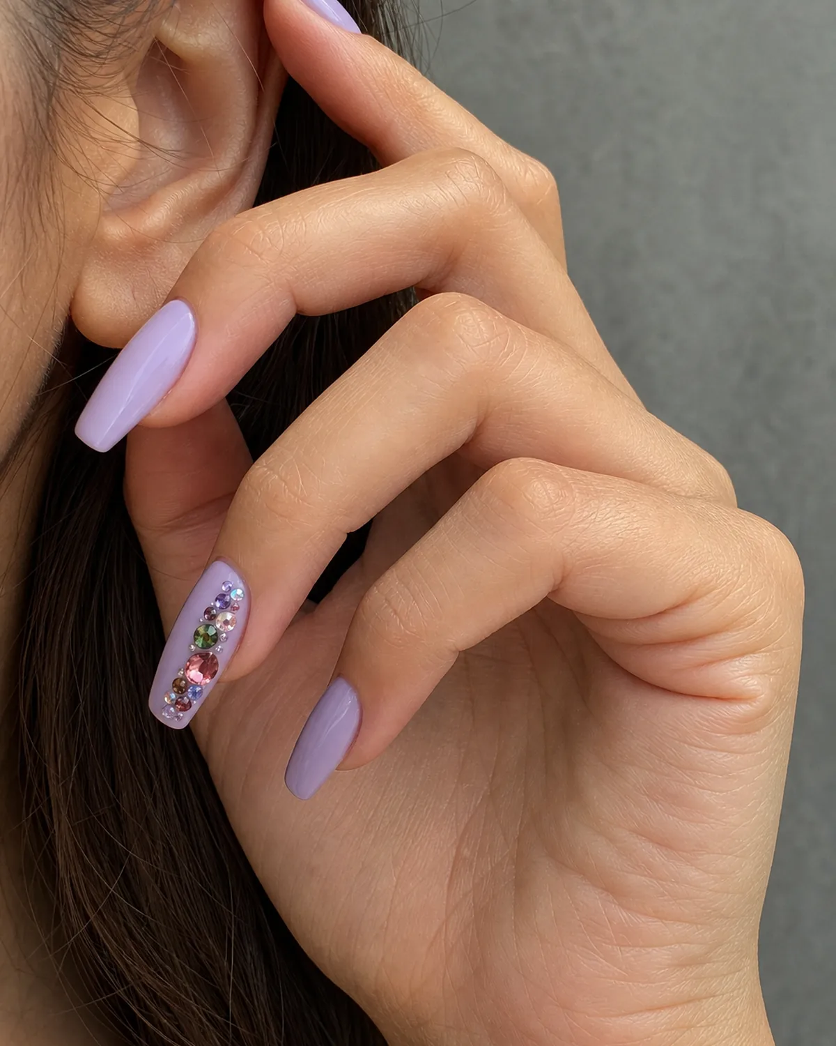 Close-up of medium coffin nails with a soft lilac base and a scattered asymmetric cluster of mixed-size raised gems, on light-medium skin tucked behind the ear