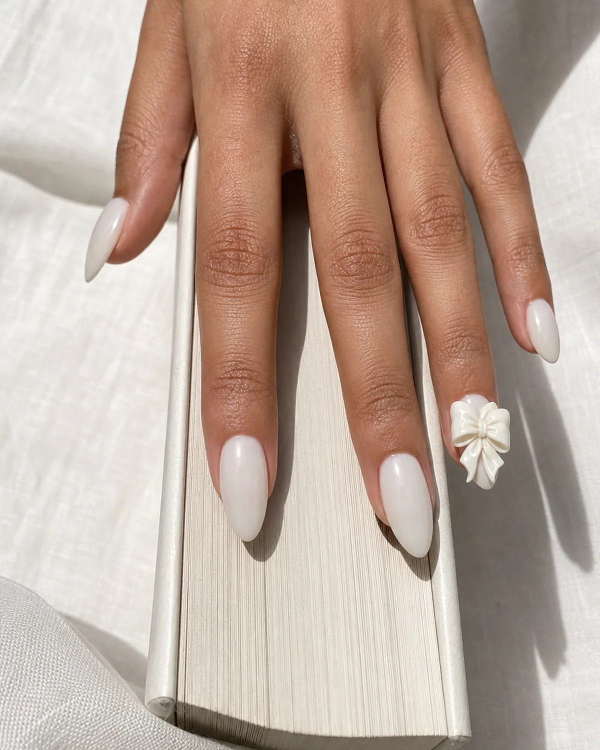 Close-up of medium almond nails with a soft ivory base and one large hand-sculpted bow with deep visible ridges, on medium skin resting on a book spine