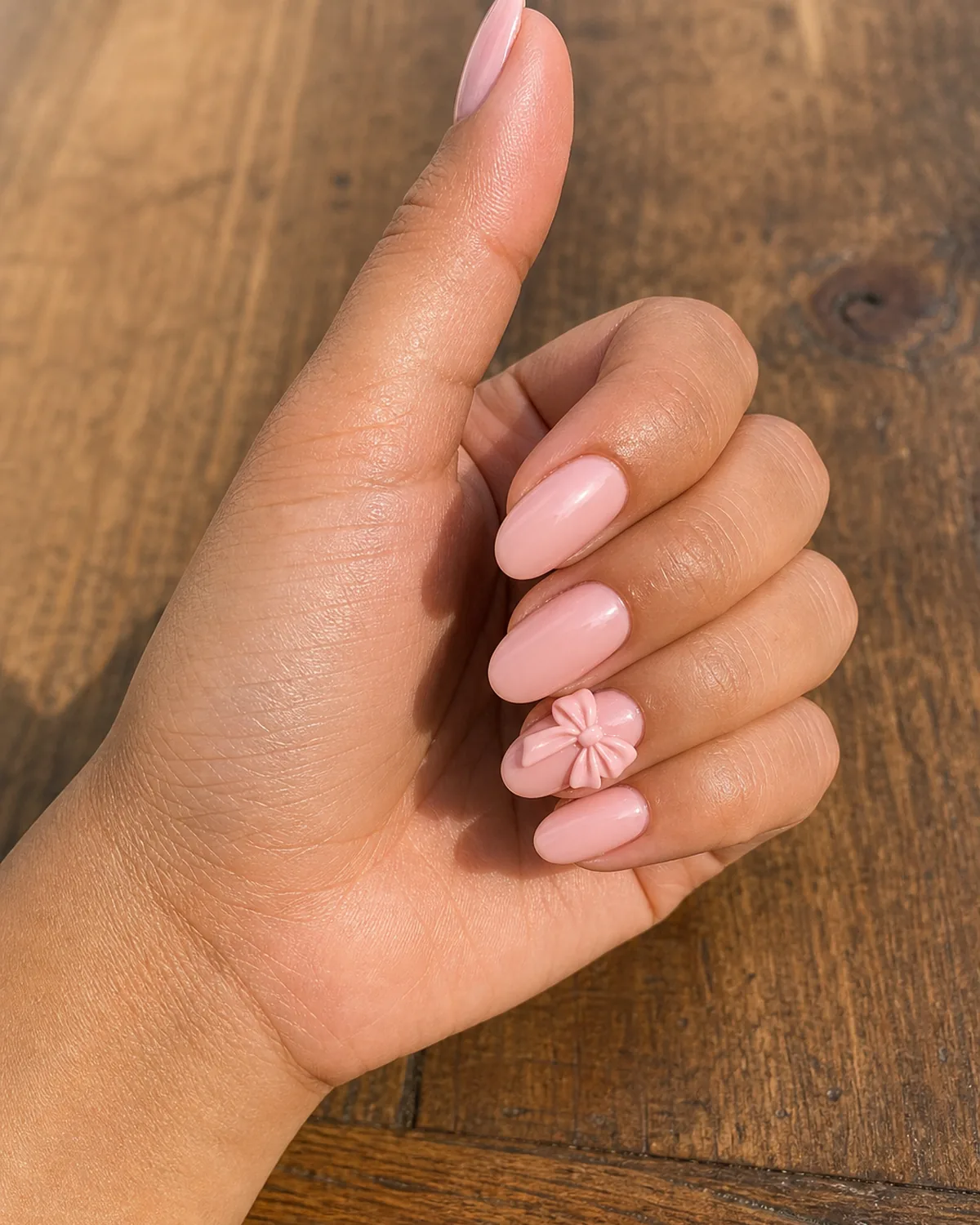 Close-up of medium oval nails with a soft pastel-pink base and a hand-sculpted acrylic bow with visible looped ridges, on tan skin turned sideways on warm walnut wood