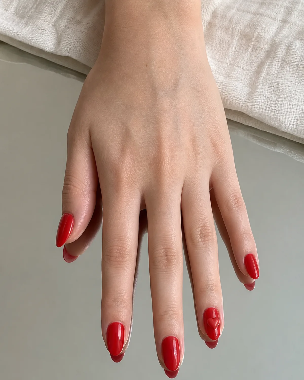 Close-up of short almond nails with a classic red base and a small sculpted gel heart sitting just barely raised above the nail plate, on light skin flat on a mirrored surface