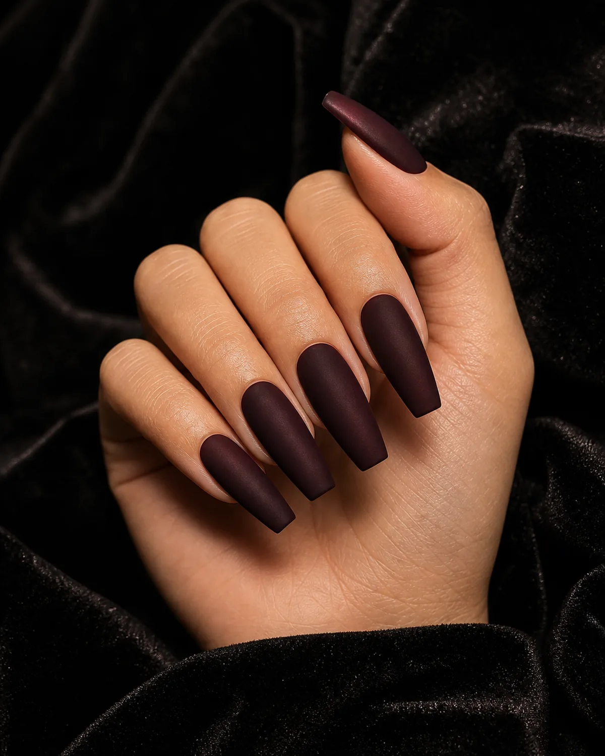 Close-up of long coffin nails in a near-black deep plum-red matte-velvet finish, on medium skin resting on black velvet fabric