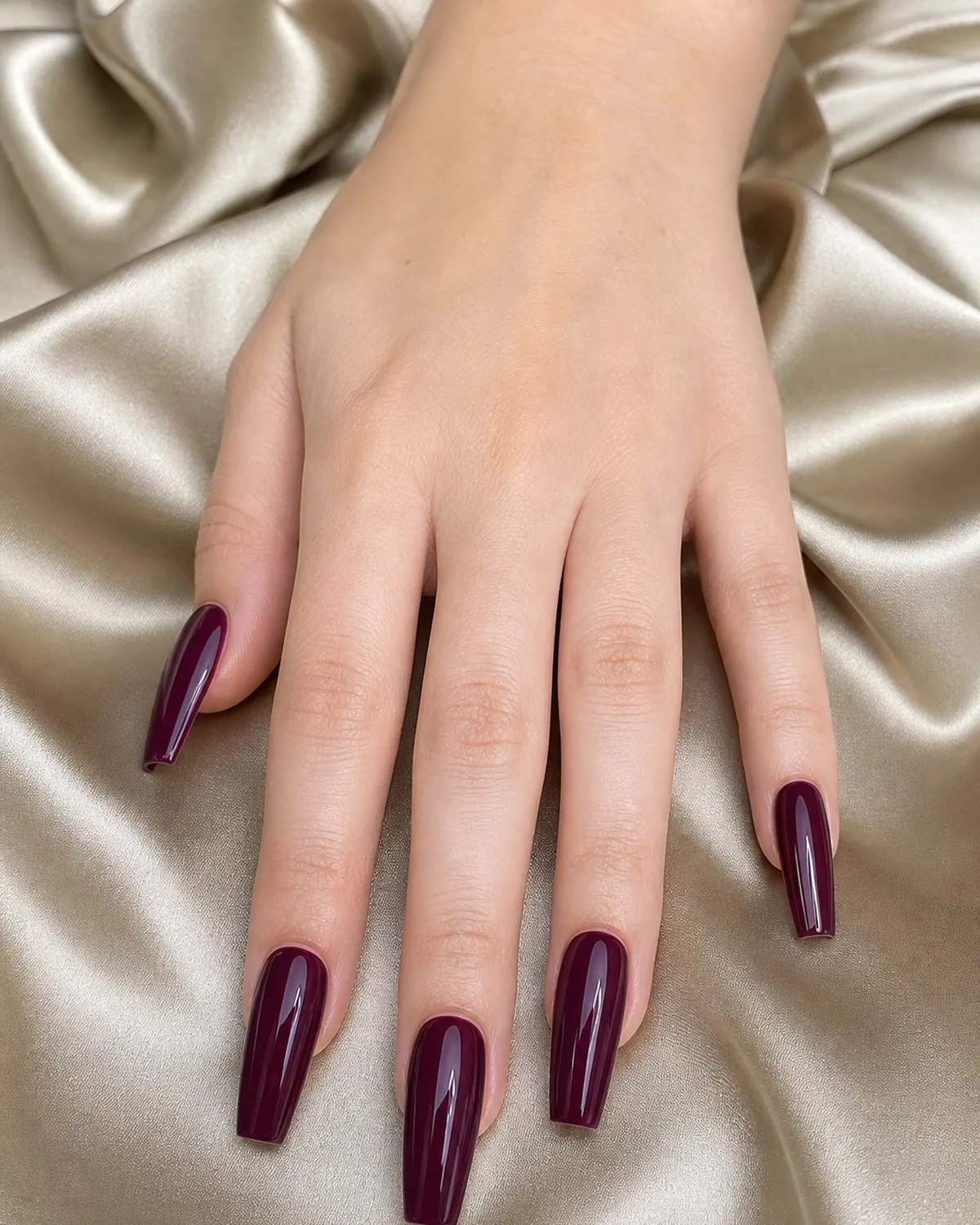 Close-up of long coffin nails in an aubergine-leaning glossy burgundy, on fair skin resting on champagne silk