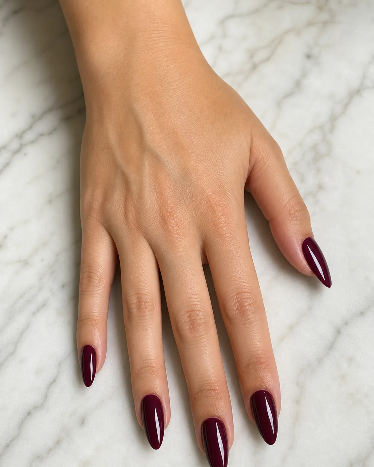 Close-up of long almond nails in a classic glossy true burgundy, full coverage, on light-medium skin resting flat on pale marble