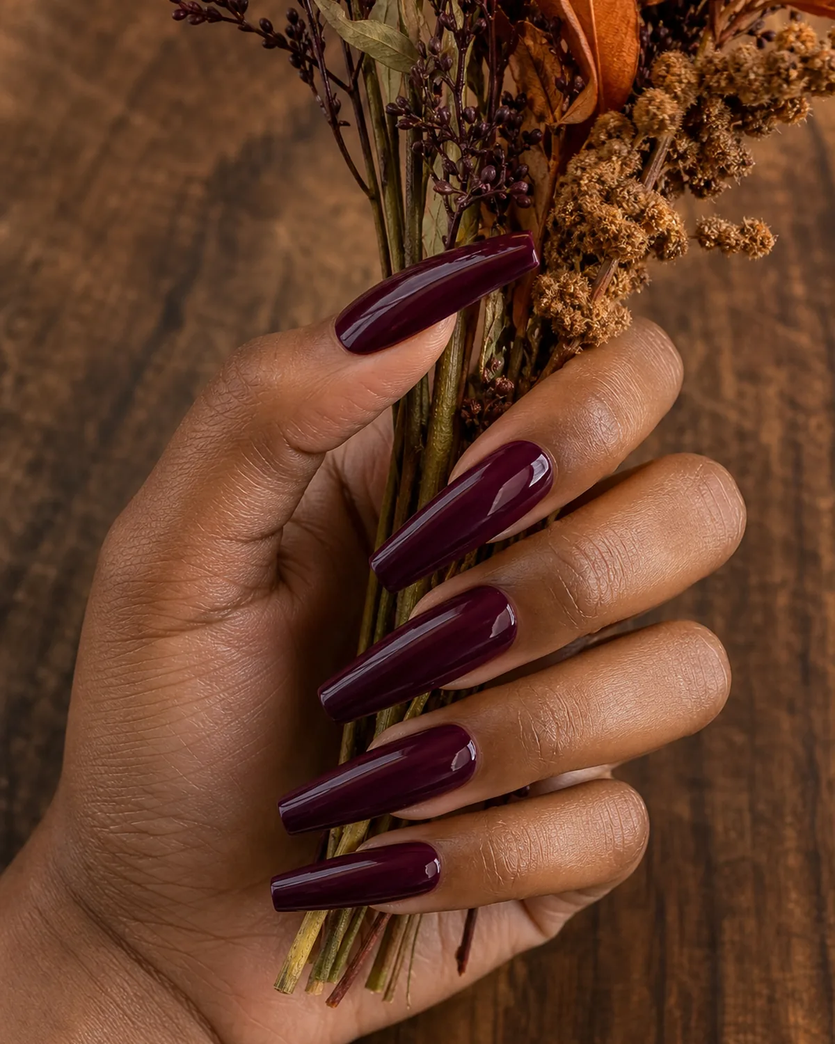 Close-up of long coffin nails in a glossy port-wine burgundy, on medium-dark skin holding a small autumn bouquet stem against warm walnut wood