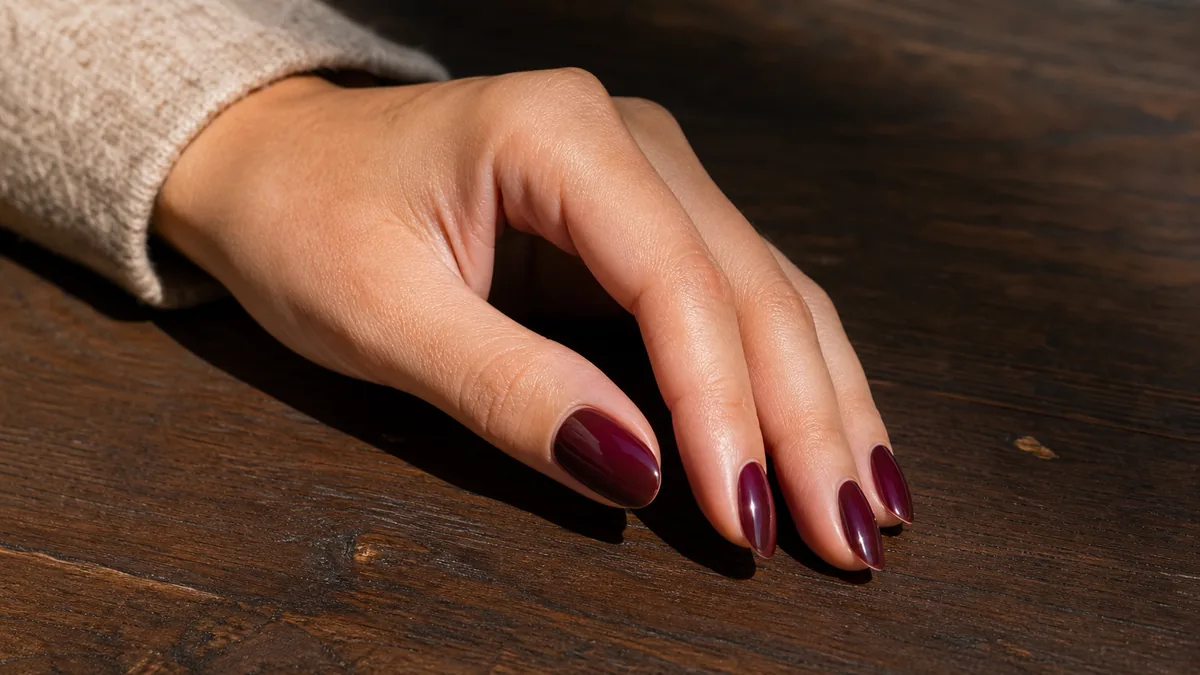 A hero close-up of one hand with medium almond nails in a classic glossy oxblood burgundy, a deep wine-red with a visible purple undertone, on light-medium skin resting on dark walnut wood