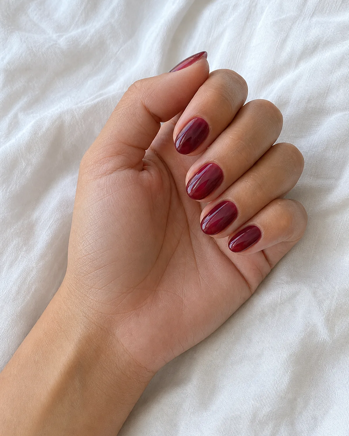 Close-up of short almond nails in a sheer garnet jelly polish with the nail bed showing faintly through, on light-medium skin resting on white linen