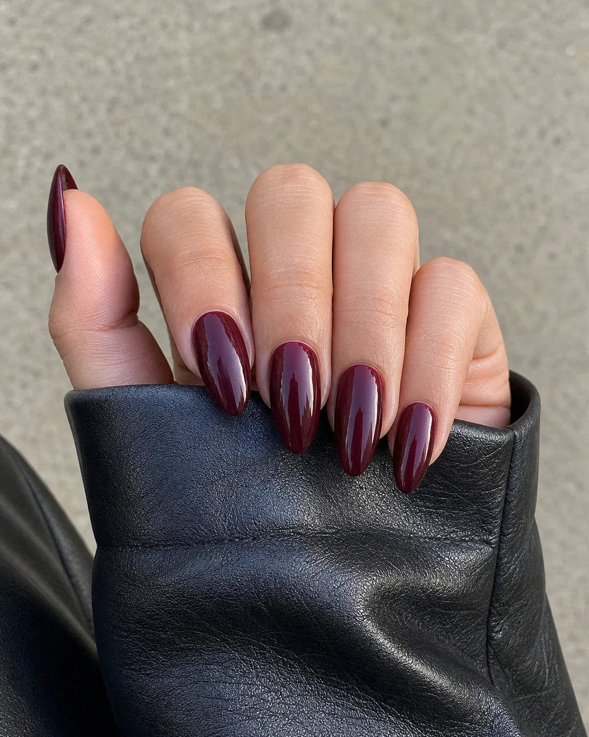 Close-up of medium almond nails in a true glossy burgundy, on light-medium skin emerging from a dark leather jacket sleeve