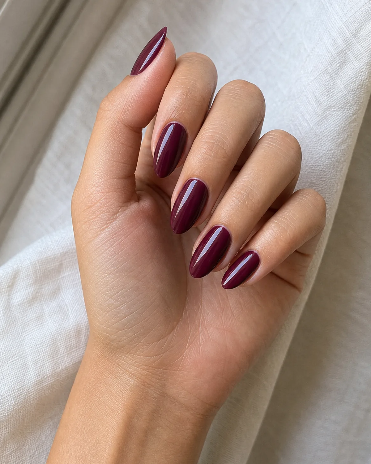 Close-up of medium almond nails in a classic glossy oxblood burgundy with a visible purple undertone, on light-medium skin turned toward a window