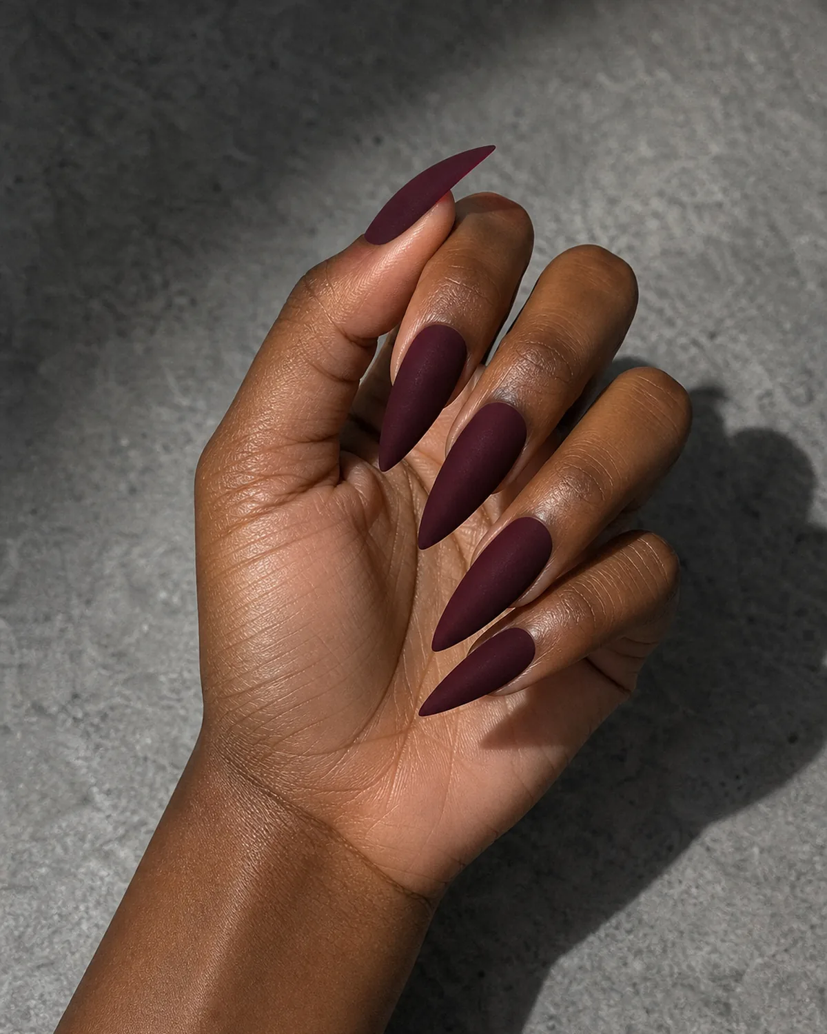 Close-up of stiletto nails in a deep wine burgundy velvet matte finish, on deep skin turned sideways in soft shadow against grey stone