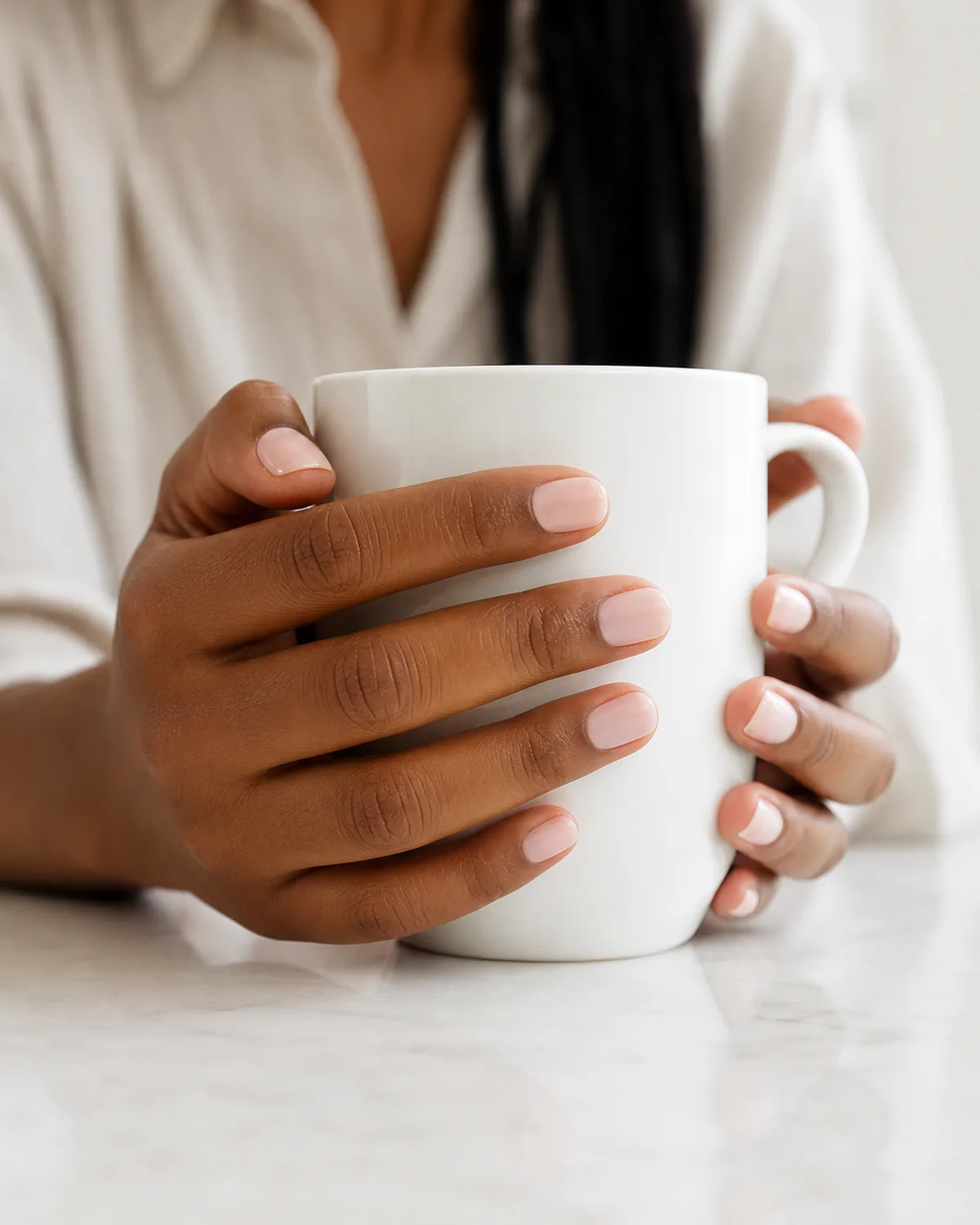Single coat soft blush on short round nails with satin finish, hand cupped around white mug
