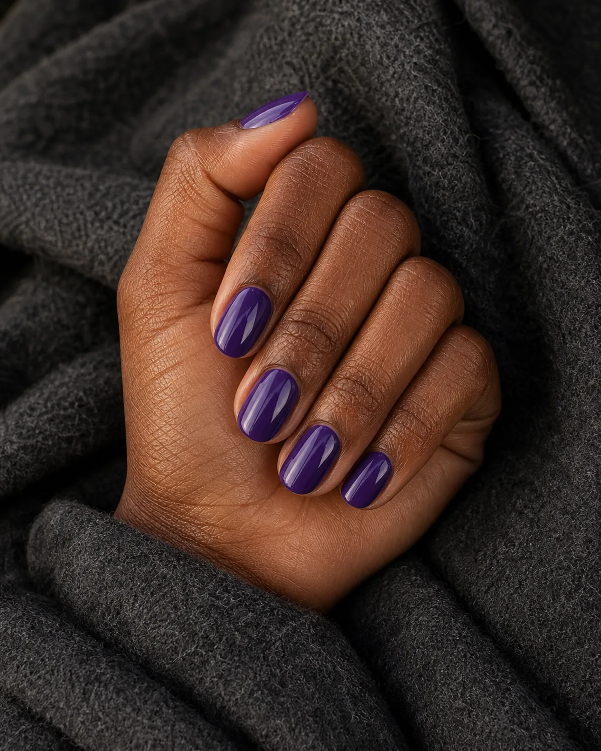 Close-up of medium round nails in a glossy grape purple, on deep skin against a charcoal wool coat