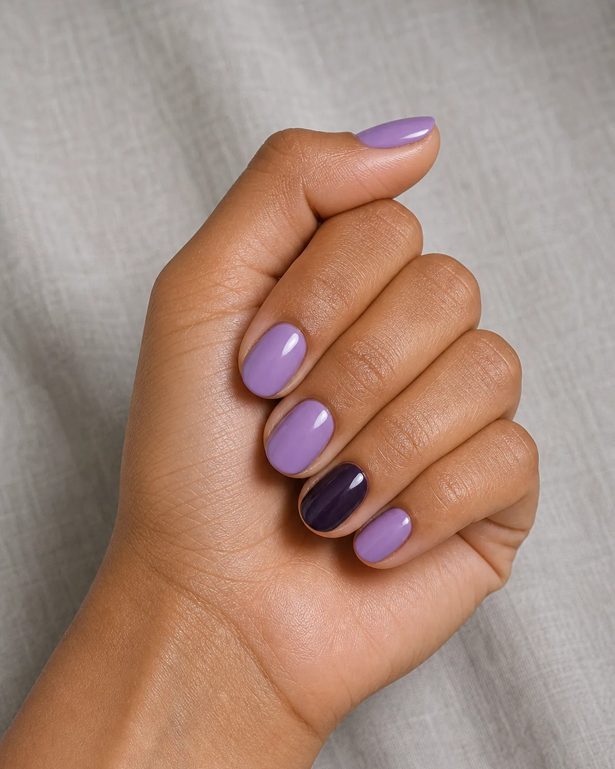 A near-black blackcurrant-purple accent nail among lavender nails on medium round shapes, a tan-skinned hand in a soft fist