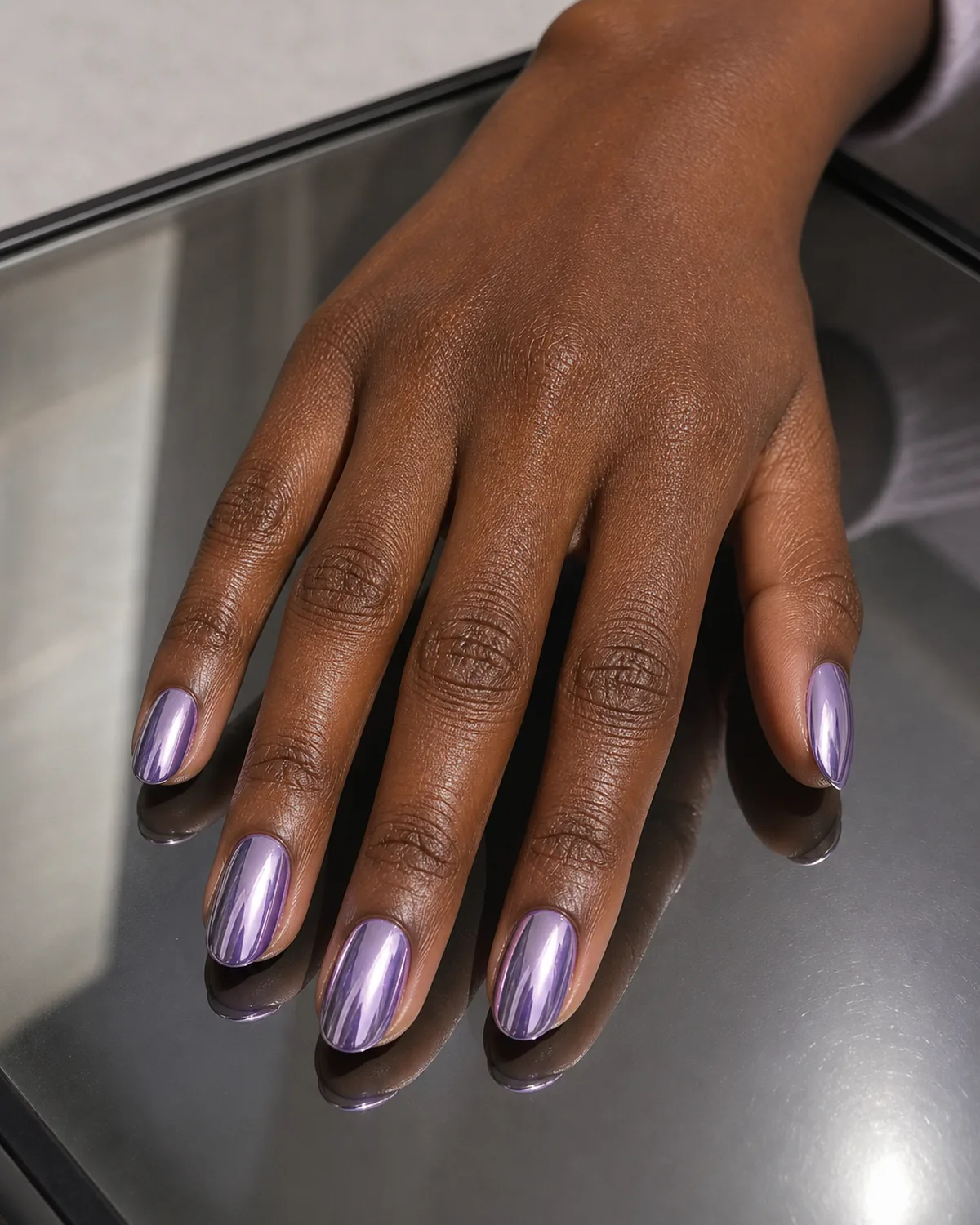 A chrome mirror finish carrying a lavender undertone on oval nails, a deep-skinned hand resting flat on a mirrored surface