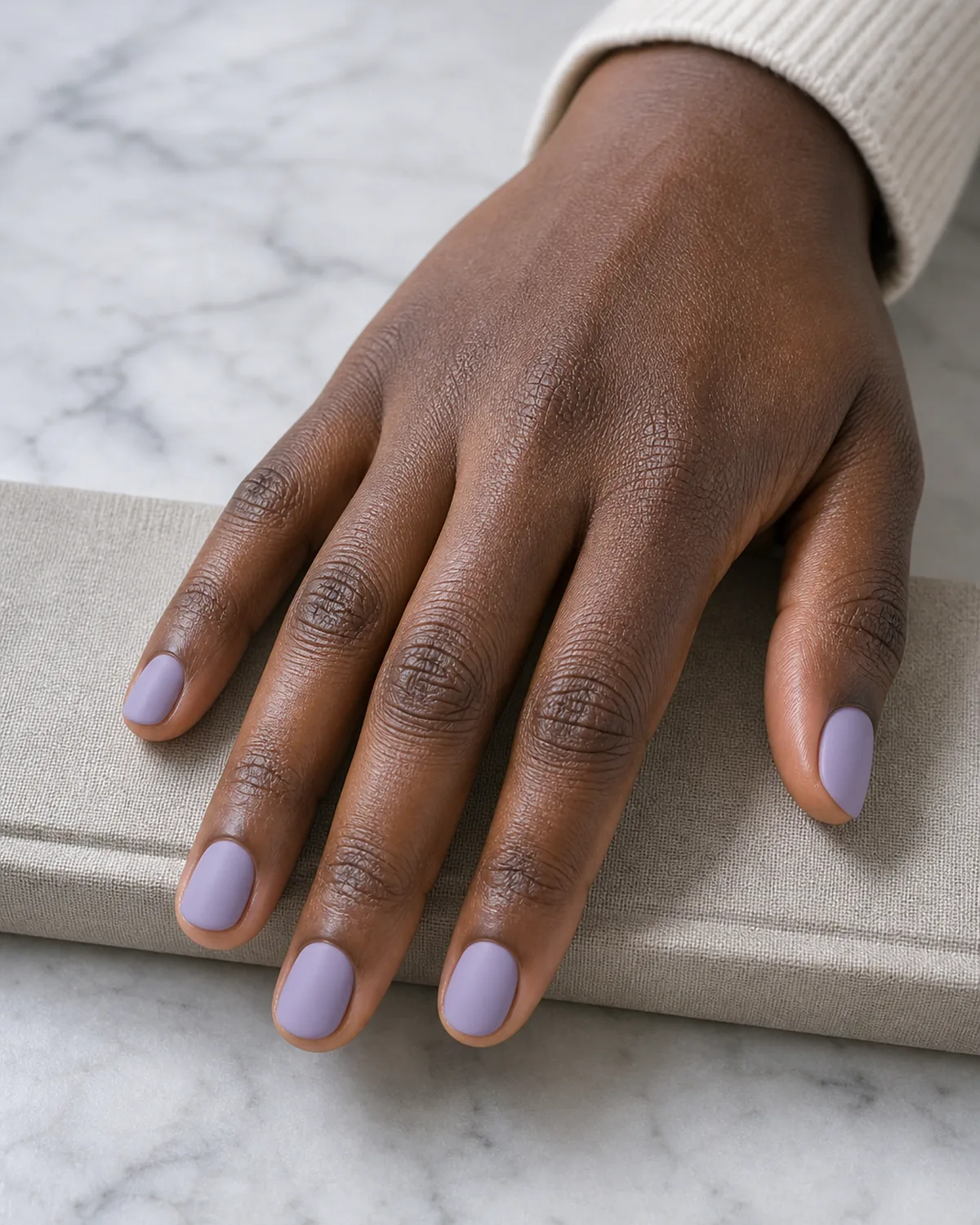 A matte lavender finish on short round nails, a deep-skinned hand resting on a linen-wrapped surface