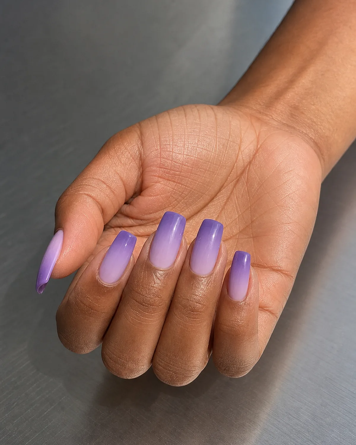 A soft ombre fading from pale lilac to deeper purple on long square nails, a medium-deep-skinned hand open palm-up
