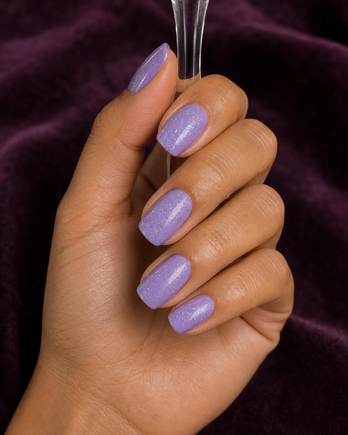 Fine silver glitter suspended in a lavender base on squoval nails, a tan-skinned hand holding a small glass stem