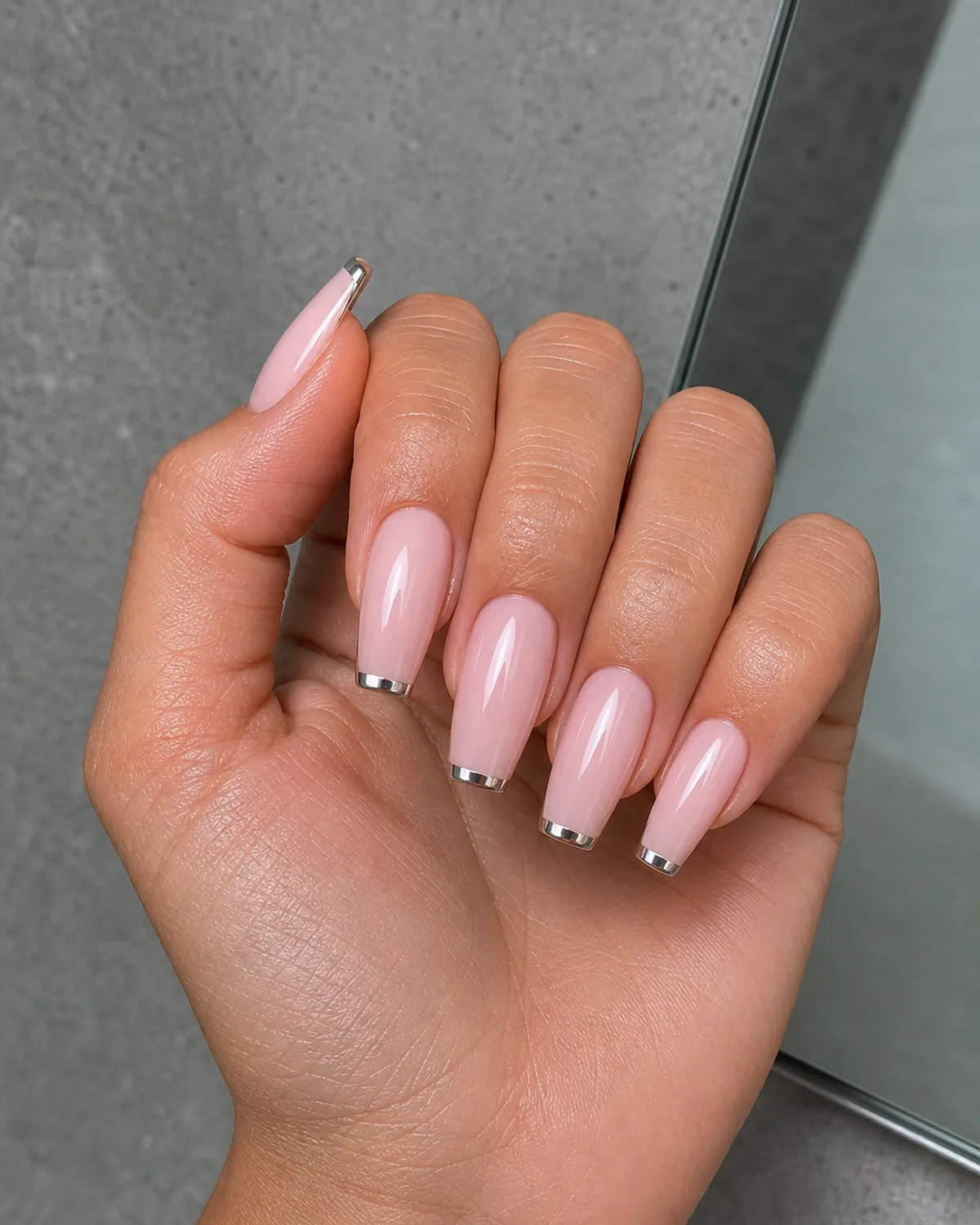 Close-up of coffin nails with a thread-thin mirrored chrome hairline tip on a pale pink wash base, on tan skin tilted toward a mirror edge