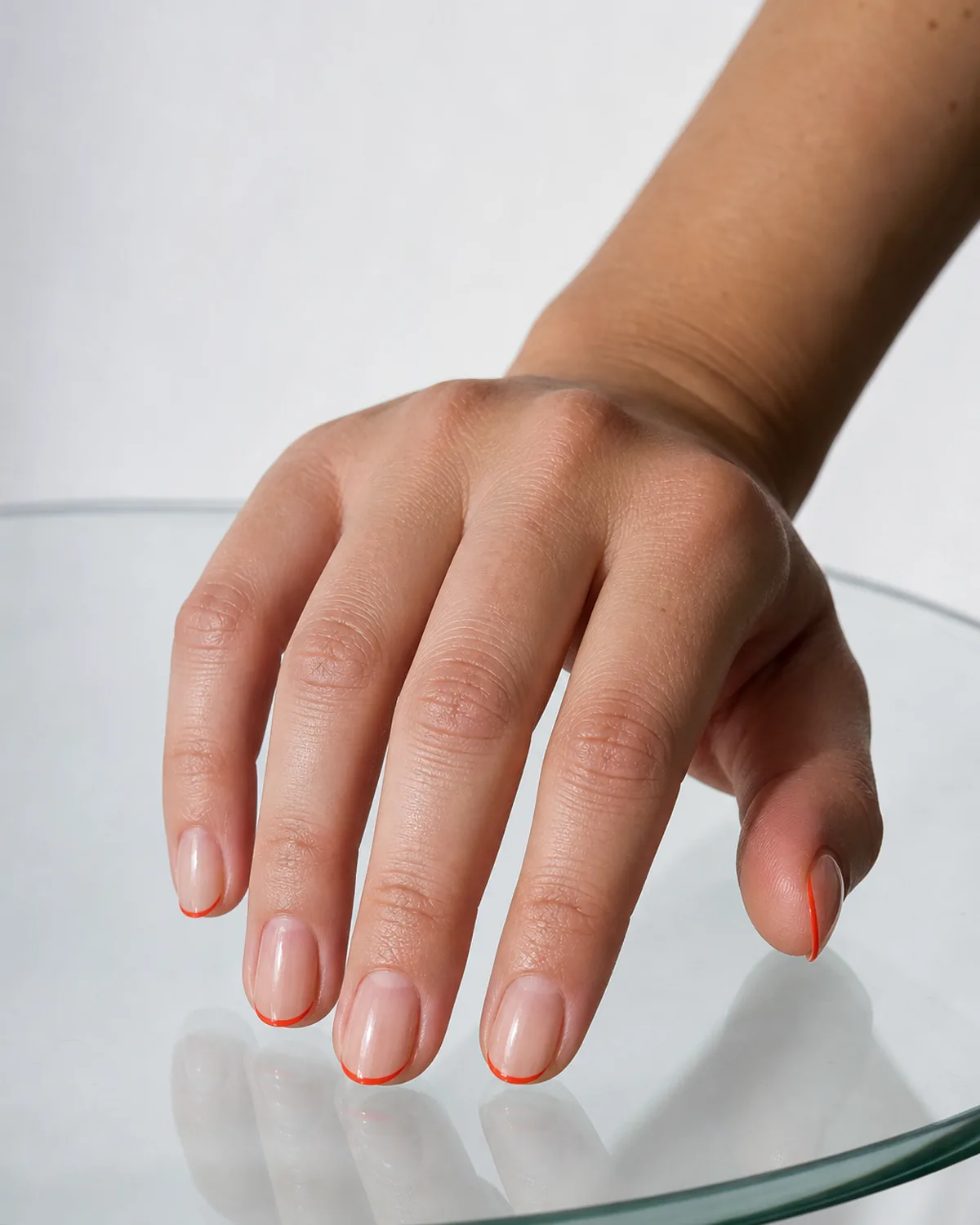 Close-up of short almond nails with a thread-thin red-orange hairline tip on a clear bare base, fingers touching a clear glass surface