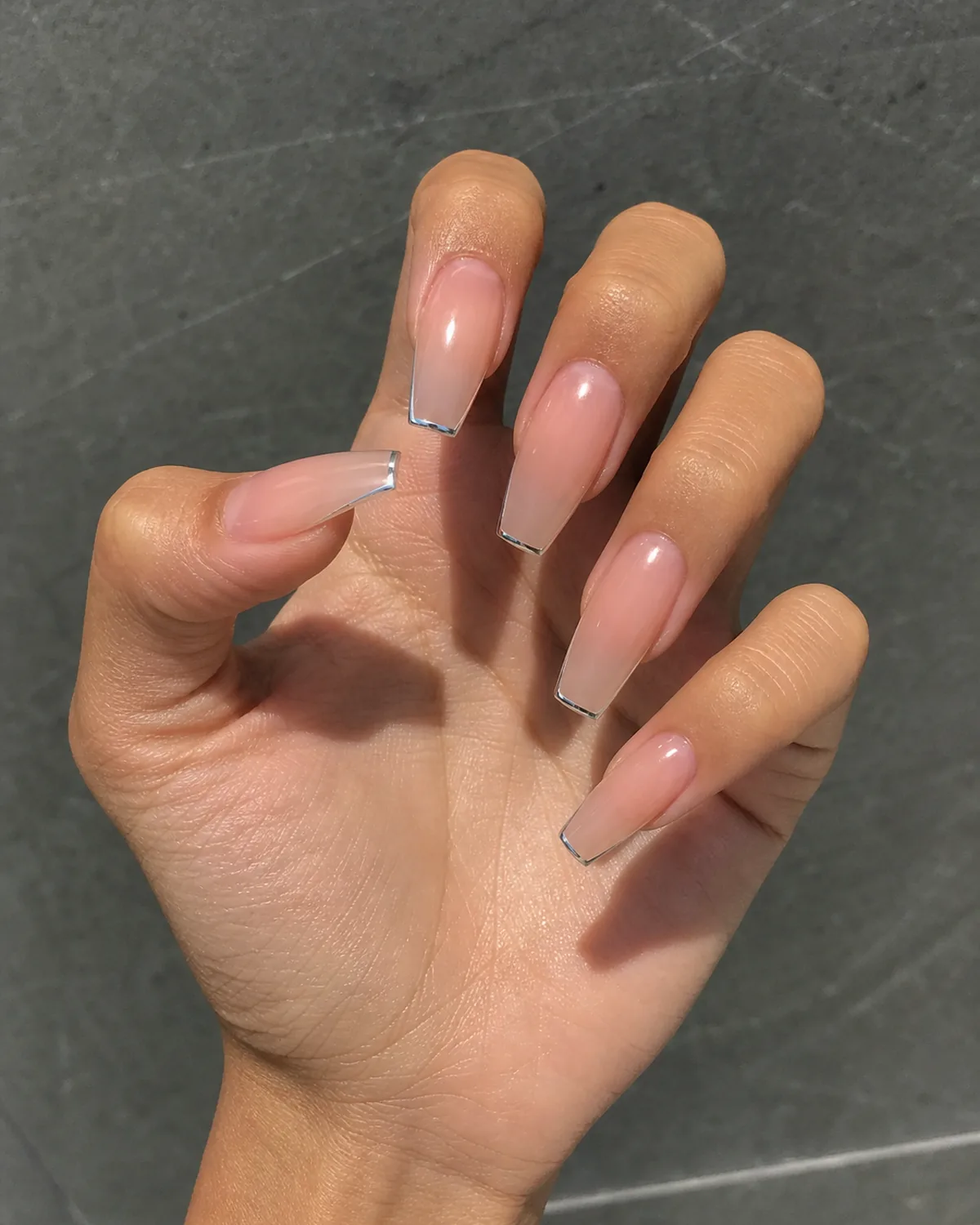 Close-up of coffin nails with a needle-thin silver chrome hairline tip on a clear bare base, on tan skin raised toward the camera