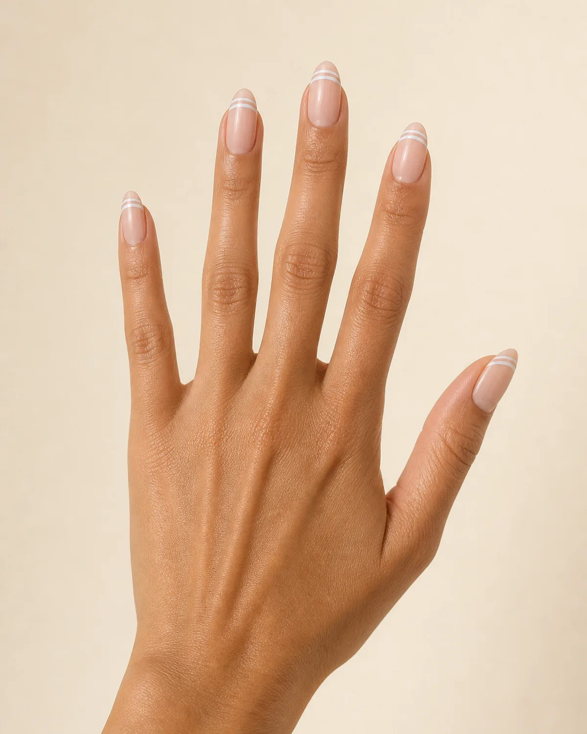 Fingers fanned against a pale blush backdrop showing almond nails with two thin parallel white lines near the tip on a nude base