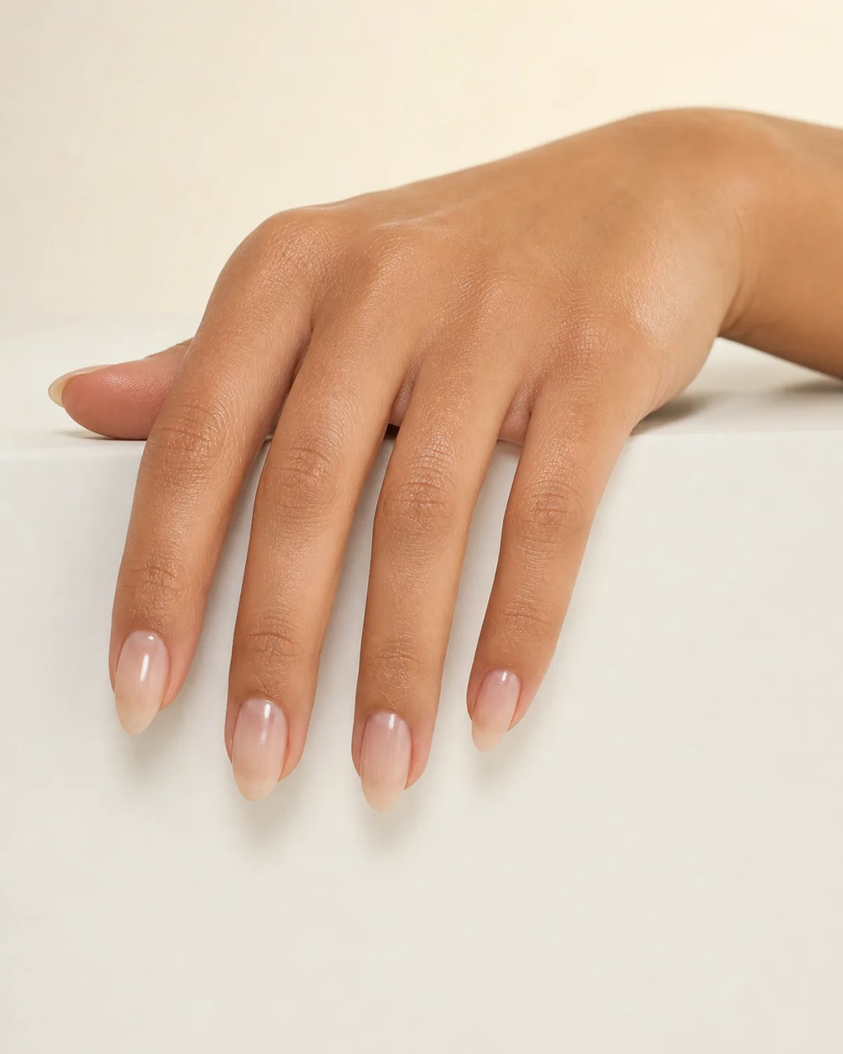 Three-quarter angle of a hand draped over a white ledge with almond nails in sheer milky gloss showing luminous shine