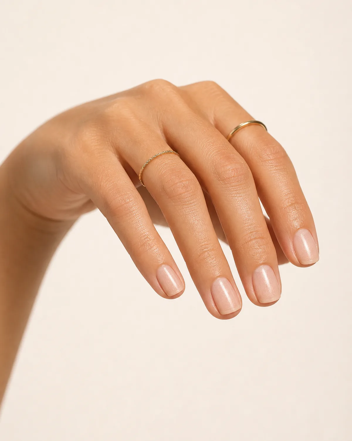 Below-angle view of a hand with short square nails in sheer nude shimmer wearing minimalist gold rings, light catching the fine shimmer