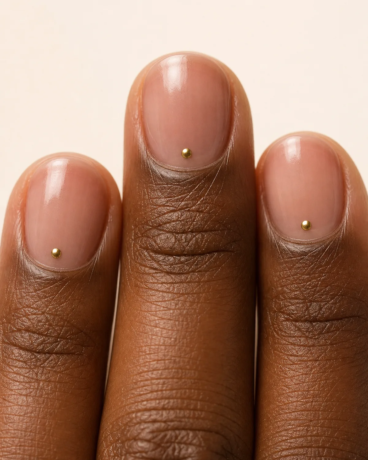Ultra-close macro of two fingertips with round nails showing a single tiny gold metallic dot near the cuticle on a bare pink wash