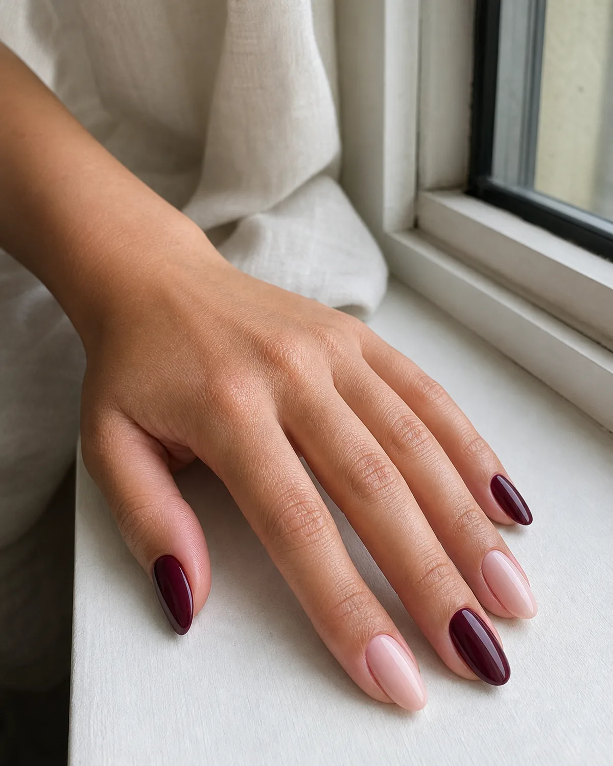 Close-up of medium almond nails alternating deep burgundy and soft blush pink nail to nail, on light-medium skin resting on a windowsill ledge