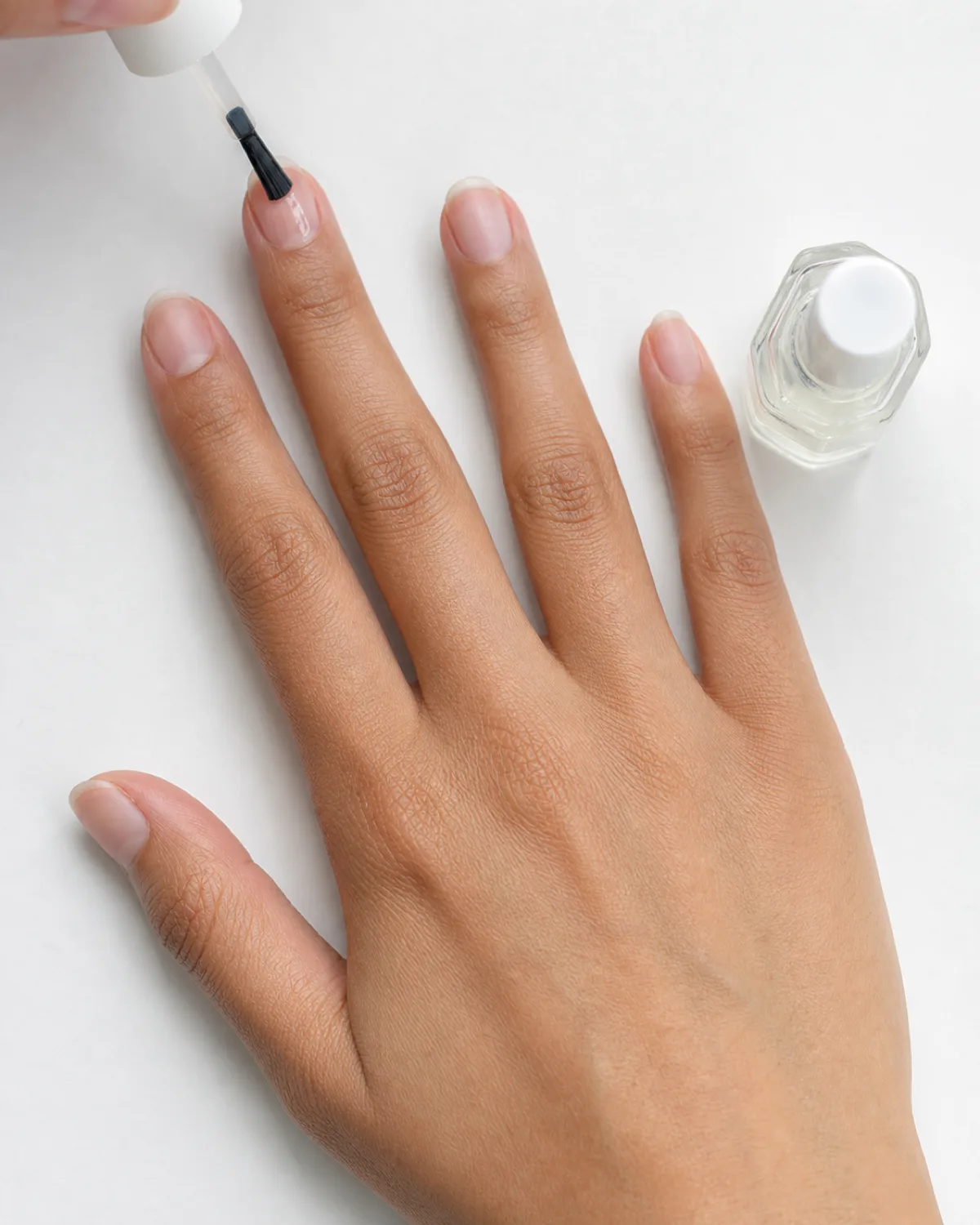 Overhead view of a hand applying clear basecoat to a natural nail with a thin brush, glossy wet coat visible