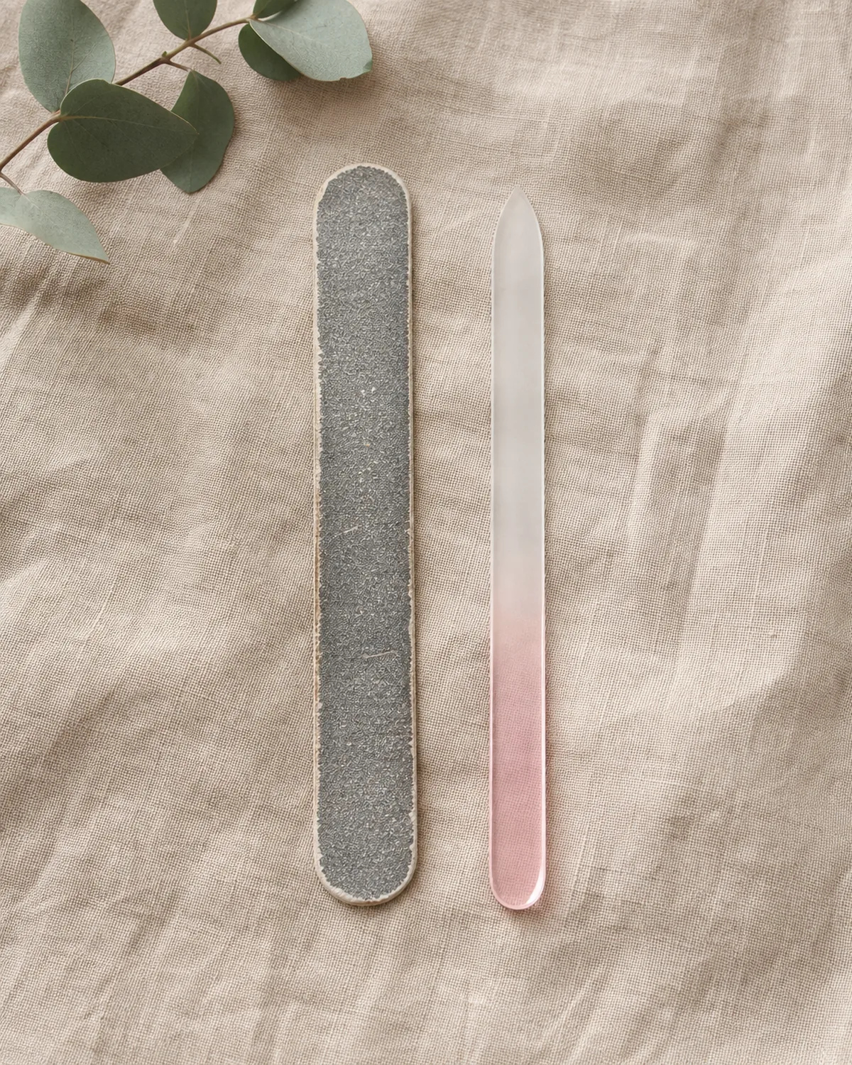 Overhead flat-lay of a crystal glass nail file beside a worn emery board on natural linen