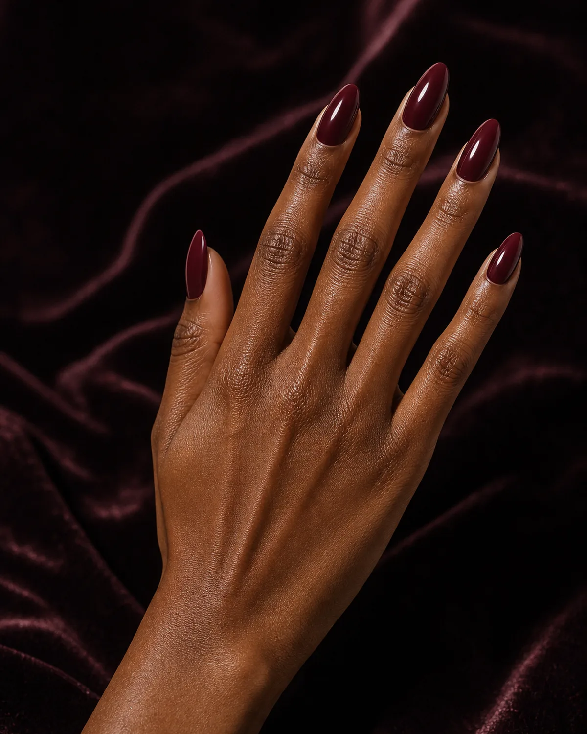 One hand with deep burgundy press-on nails on almond shape against dark textured fabric, side profile angle with fingers reaching up in an elegant gesture