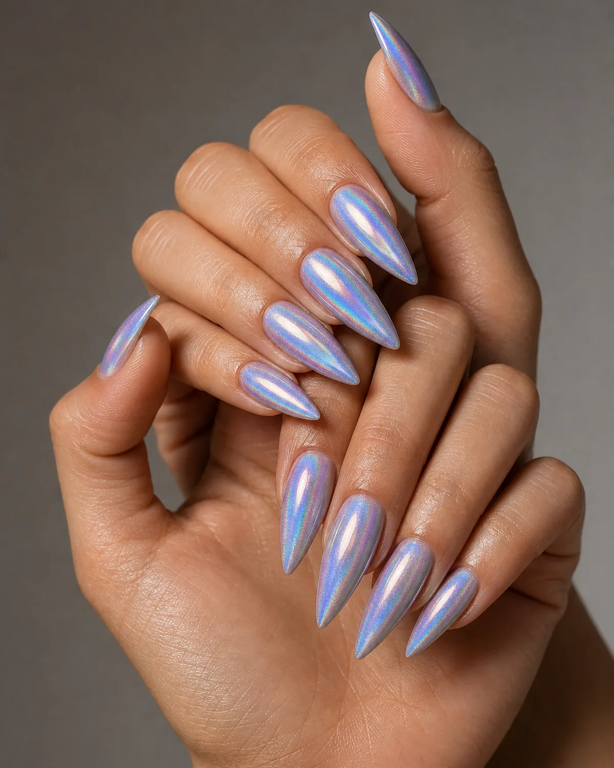 Three-quarter angle of hands with long stiletto press-on nails in holographic iridescent finish, dramatic pointed length, fingers loosely interlaced, light catching the holographic shift