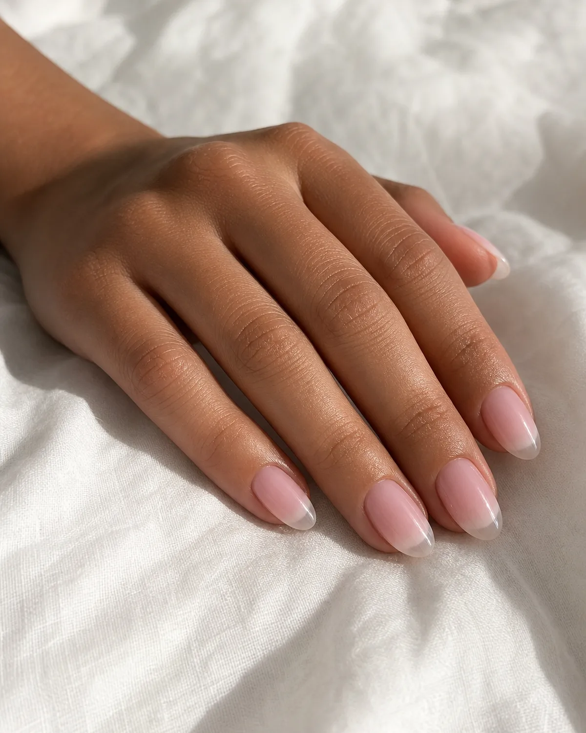 Side angle of a hand with short almond acrylic nails showing baby pink ombré fading to a clear tip on white linen